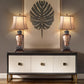 Two table lamps on a console table with a decorative wall piece in the background.
