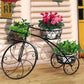 Black metal plant stand with three tiers holding potted plants against a wooden wall.