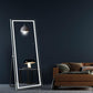 Large illuminated standing mirror in a room with a dark wall and brown sofa.