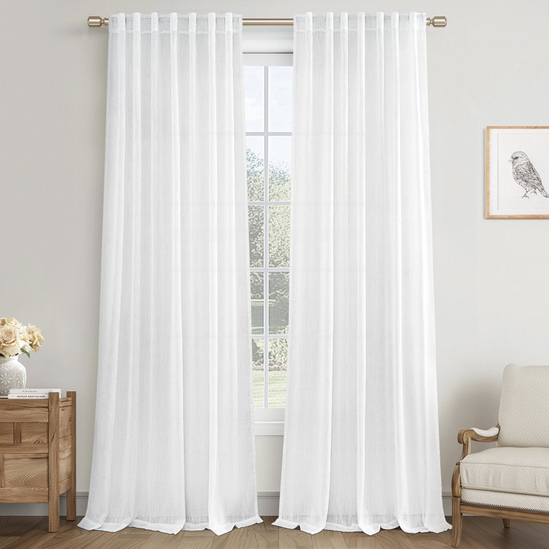 White sheer curtains hanging in front of a window with a side table and chair in the background.