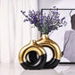 Two gold and black vases with purple flowers on a white surface.