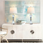 Two table lamps with crystal bases on a white console table in front of abstract art.