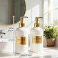 Two white soap dispensers with gold labels on a bathroom counter, next to a plant and mirror.