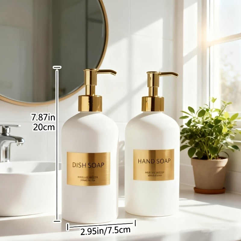 Two white soap dispensers with gold labels on a bathroom counter, next to a plant and mirror.