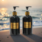 Two black soap dispensers with gold labels on a beach at sunset.