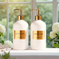 Two white soap dispensers with gold labels on a windowsill with flowers in the background