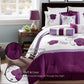 Purple and white floral bedding set in a bedroom with a lamp and plant.