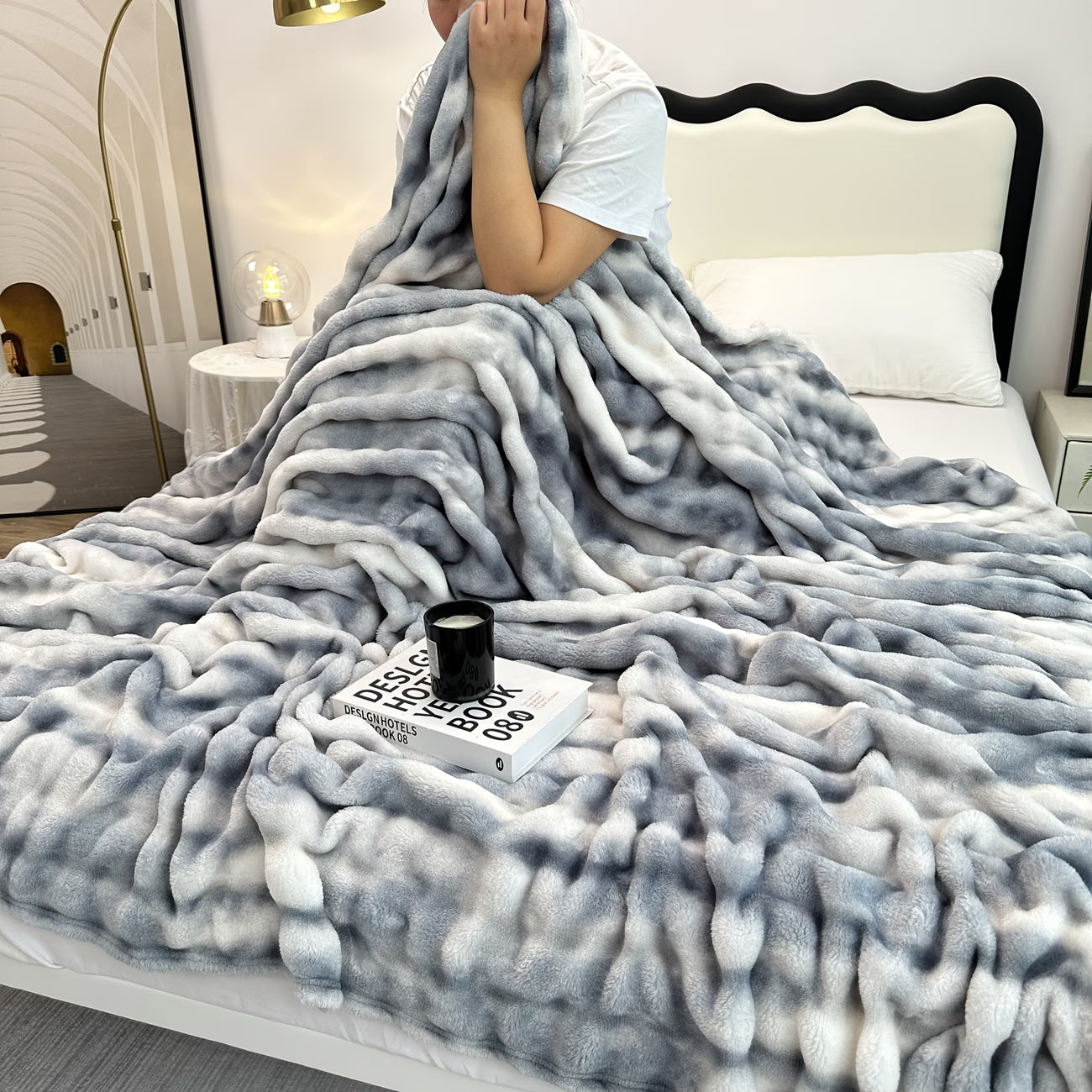 Person wrapped in a fluffy gray blanket on a bed with a book and mug beside them.