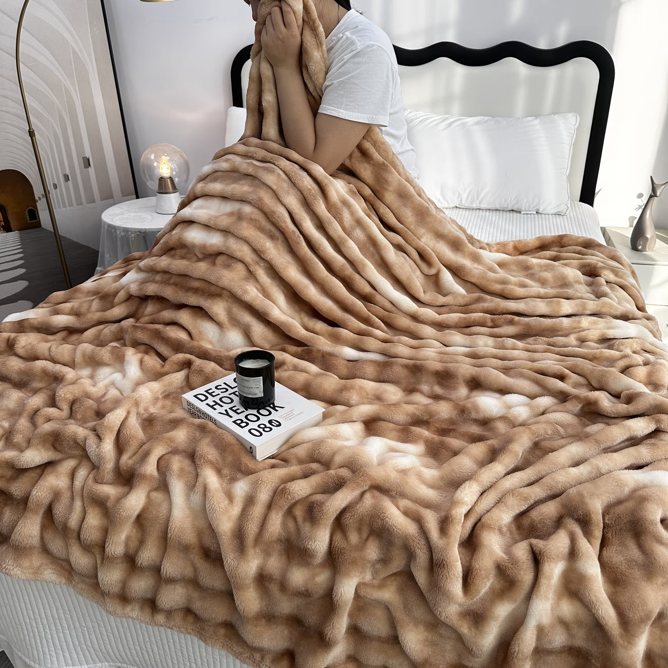 Person wrapped in a fluffy beige blanket on a bed with a candle and book on the nightstand.