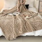 Beige textured blanket draped over a couch with magazines and a candle on a white surface.