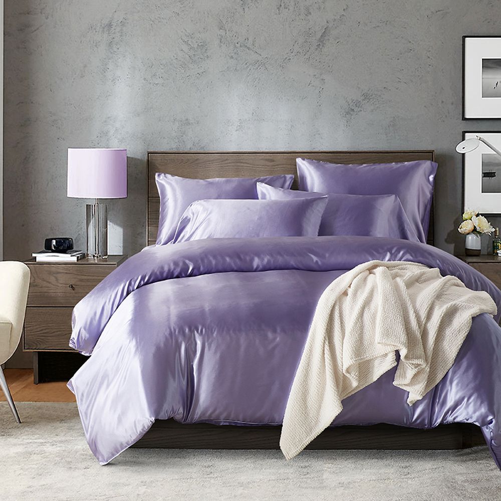 Purple satin bedding set on a bed in a room with a gray wall and a lamp.