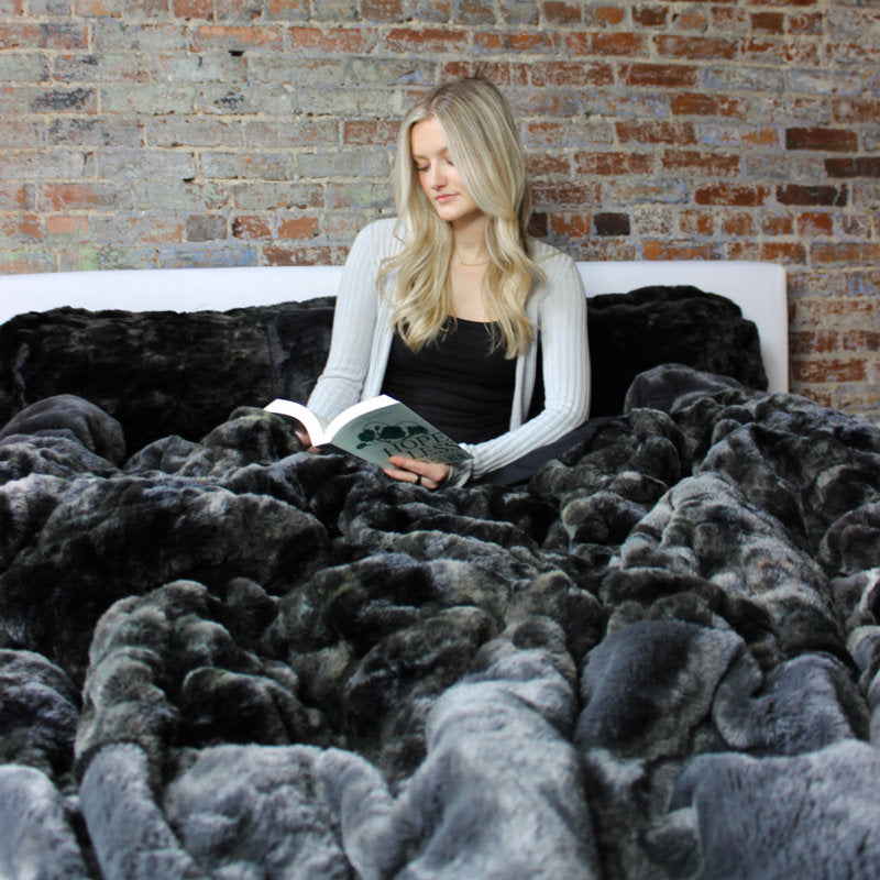 Woman sitting on a bed with a large gray blanket, reading a book against a brick wall.