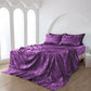 Purple patterned bedspread and pillows on a bed in a bright room.