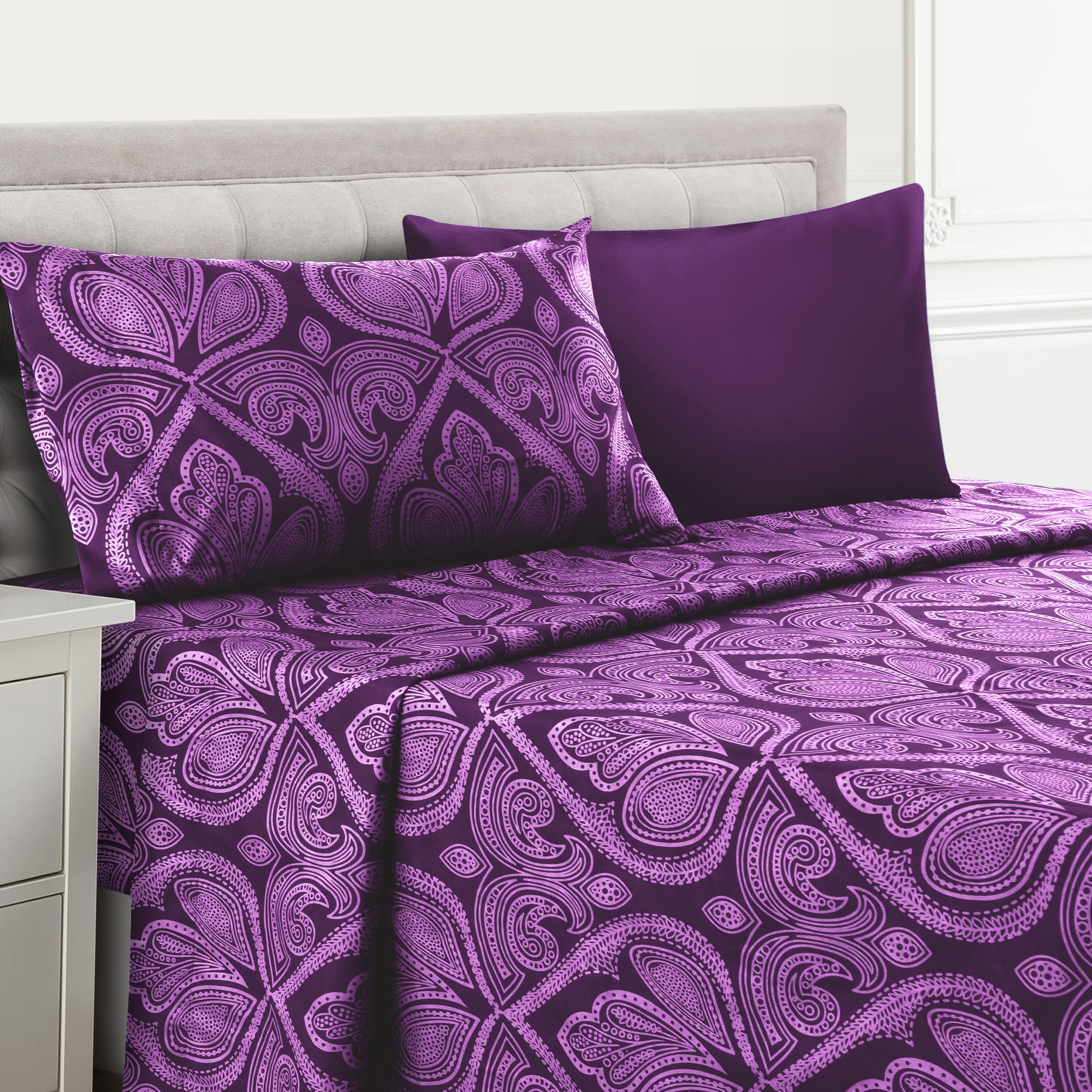 Purple paisley patterned bedding set on a bed with a white headboard.