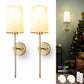 Pair of gold wall sconces with white lampshades on a white background, with a Christmas tree and presents in the background.