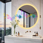 Bathroom vanity with a round illuminated mirror, decorative elements, and a view of trees outside.