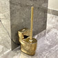 Gold toilet brush holder with brush against a marble bathroom wall.