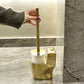 Gold toilet brush holder with a hand holding the handle in a bathroom setting.