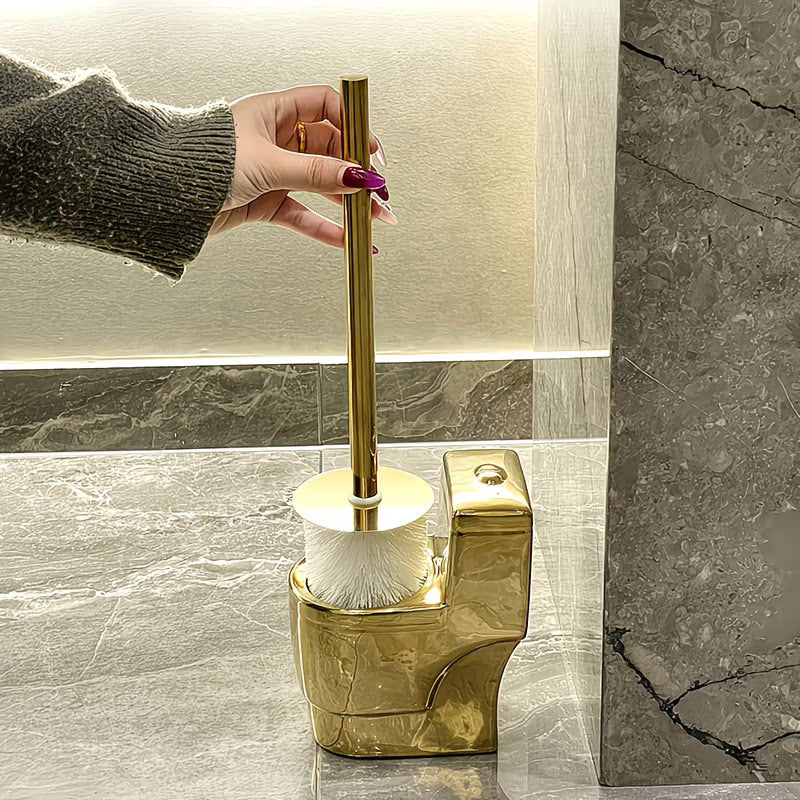 Gold toilet brush holder with a hand holding the handle in a bathroom setting.