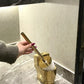 Gold toilet brush holder with a hand holding a brush against a marble bathroom wall.
