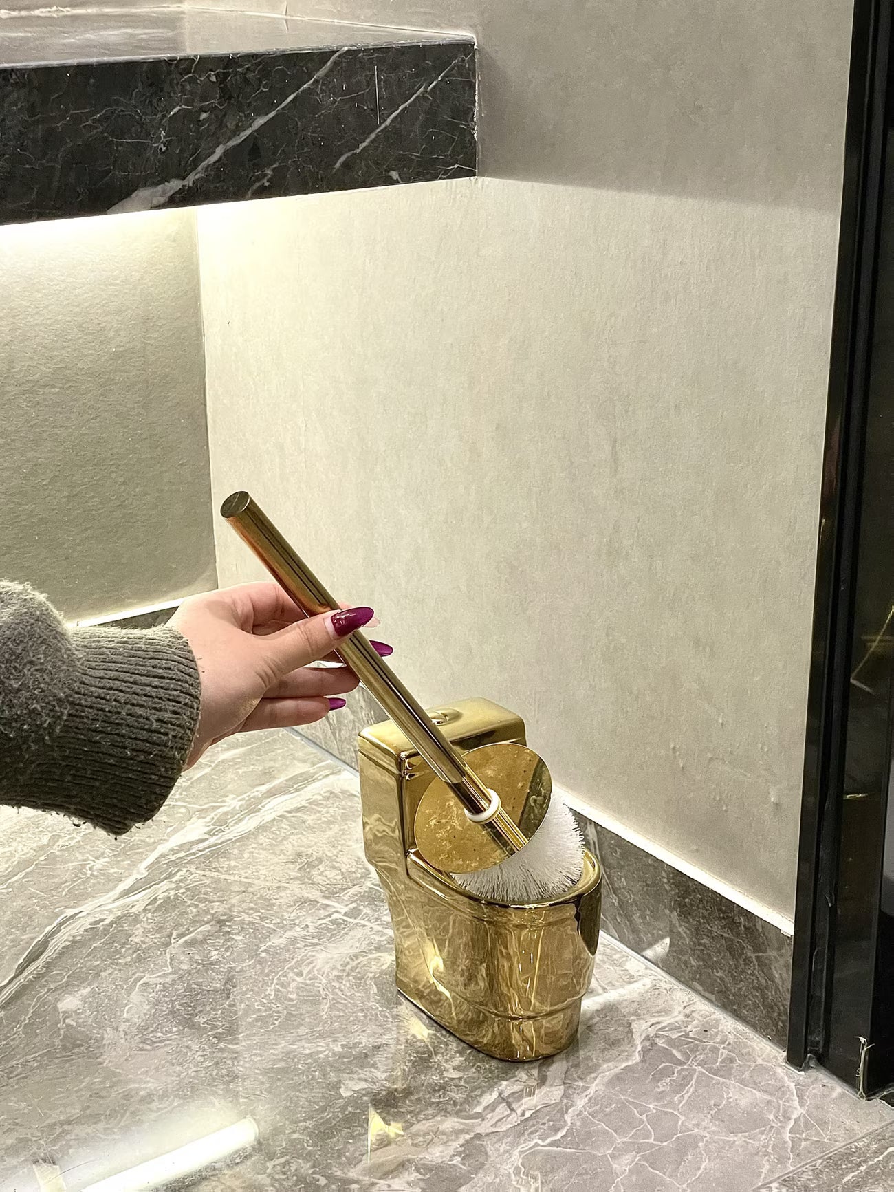 Gold toilet brush holder with a hand holding a brush against a marble bathroom wall.