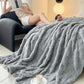 Gray textured blanket draped over a couch with a person reading a book in the background.