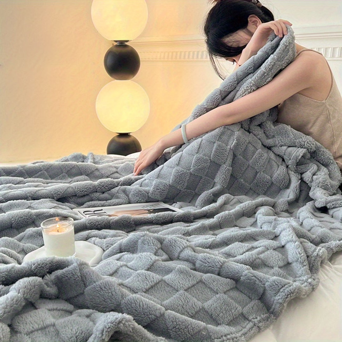 Person wrapped in a soft gray blanket in a cozy room with a lamp and candle.