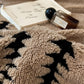 Close-up of a textured beige blanket with black patterns, a book, and a bottle on a surface.