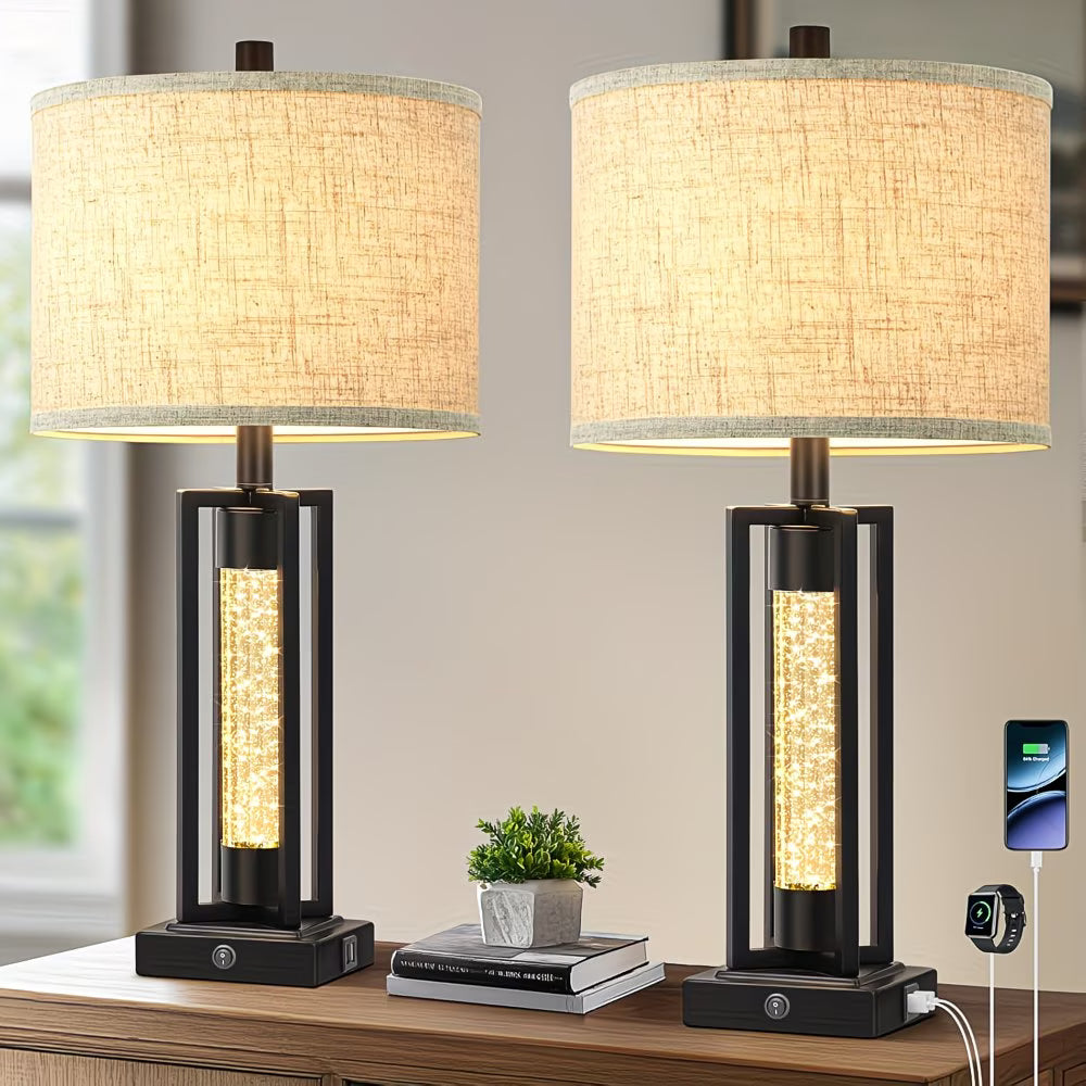 Two table lamps with fabric shades on a wooden surface, with a smartphone and small plant in the background.